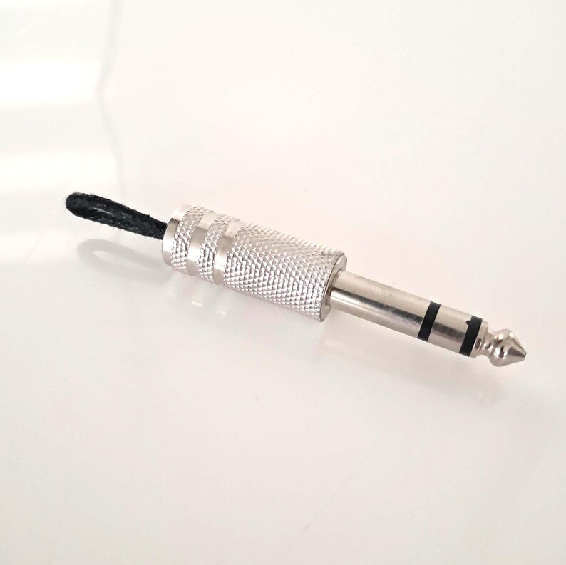 May include: A silver-colored 1/4 inch audio connector with a textured grip and a black cord loop. The connector has a tip, two black bands, and a silver shaft. The background is white.