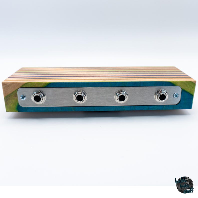 May include: A wooden audio device with four input jacks. The device has a rectangular shape with a multi-colored striped top and a teal blue front. A silver metal plate holds the input jacks.