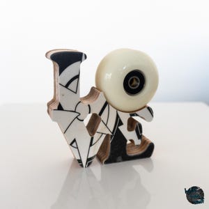 Puede incluir: Una escultura decorativa de madera que deletrea "LOVE" con una rueda de monopatín. Las letras son negras y blancas con patrones geométricos. La rueda del monopatín es blanquecina. La escultura está sobre una superficie blanca.