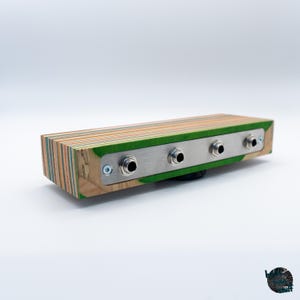 May include: A rectangular wooden audio effects pedal with a colorful, layered design. The pedal features a brushed metal plate with four input jacks. The wood has green, orange, and natural wood tones.