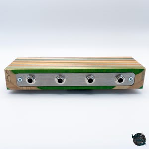 May include: A rectangular audio device with a wooden body made from layered skateboard decks. The front features a brushed metal plate with four input jacks. The wood has green, yellow, and orange stripes.