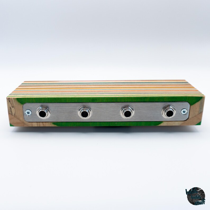 May include: A rectangular audio device with a wooden body made from layered skateboard decks. The front features a brushed metal plate with four input jacks. The wood has green, yellow, and orange stripes.