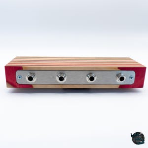 May include: A rectangular wooden audio device with four input jacks. The top is made of layered wood in shades of red, tan, and orange. The front panel is brushed metal with four black input jacks. The sides are red.