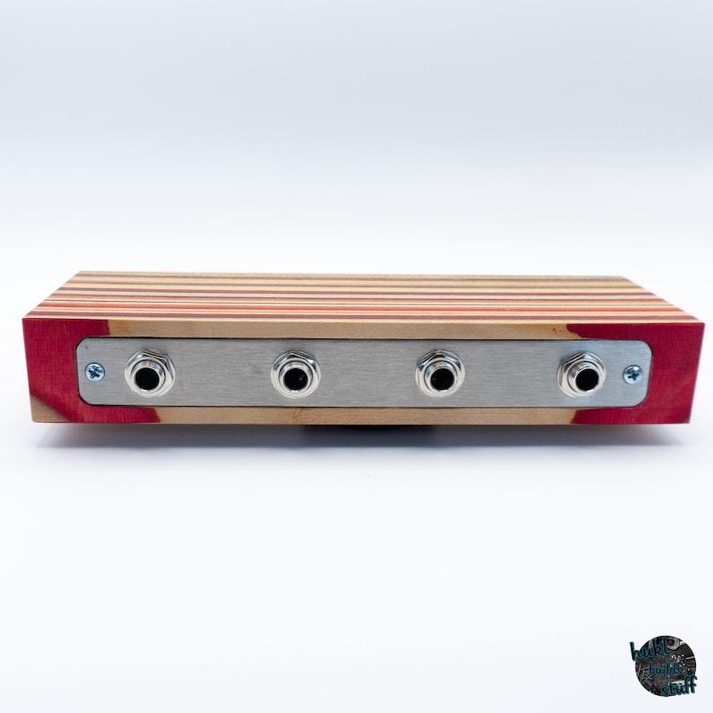 May include: A rectangular wooden audio device with four input jacks. The top is made of layered wood in shades of red, tan, and orange. The front panel is brushed metal with four black input jacks. The sides are red.