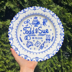 Pode incluir: Um prato de cerâmica branca com desenhos intrincados pintados à mão em azul. O centro apresenta o texto Todd & Sue, a data 08.18.2026 e ilustrações de um bolo de casamento, anéis, taças de champanhe, um carro e um envelope.