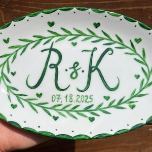 Puede incluir: Plato ovalado de cerámica blanca con borde festoneado verde y diseño floral verde. El plato presenta las iniciales "R & K" y la fecha "07.18.2025" en escritura verde, rodeadas por una corona verde y detalles de corazón.