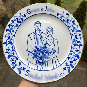 Puede incluir: Un plato de cerámica blanco con un borde azul y dorado. El plato presenta una ilustración pintada a mano de una pareja en un entorno de boda. La pareja sostiene un ramo de flores. El plato está inscrito con el texto "G.G. & J.S. 2024".