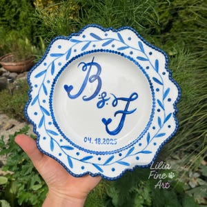 Puede incluir: Plato de cerámica blanco con borde festoneado, adornado con motivos florales azules y un borde punteado azul. El plato presenta las iniciales "B & F" y la fecha "04.18.2025" en escritura azul. Firmado "Lilia Fine Art".
