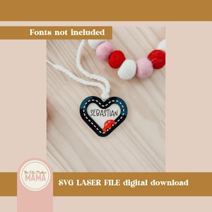 May include: A black heart-shaped wooden ornament with a white rope attached. The ornament has a white border and the name "Sebastian" written inside. A red car is also inside the heart. The ornament is on a wooden surface.
