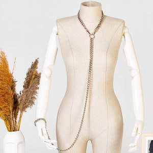 Può includere: Un set di collana e braccialetto a catena argentata con un anello sulla collana. La catena è lunga e arriva fino alla vita. Il set è esposto su un manichino bianco.