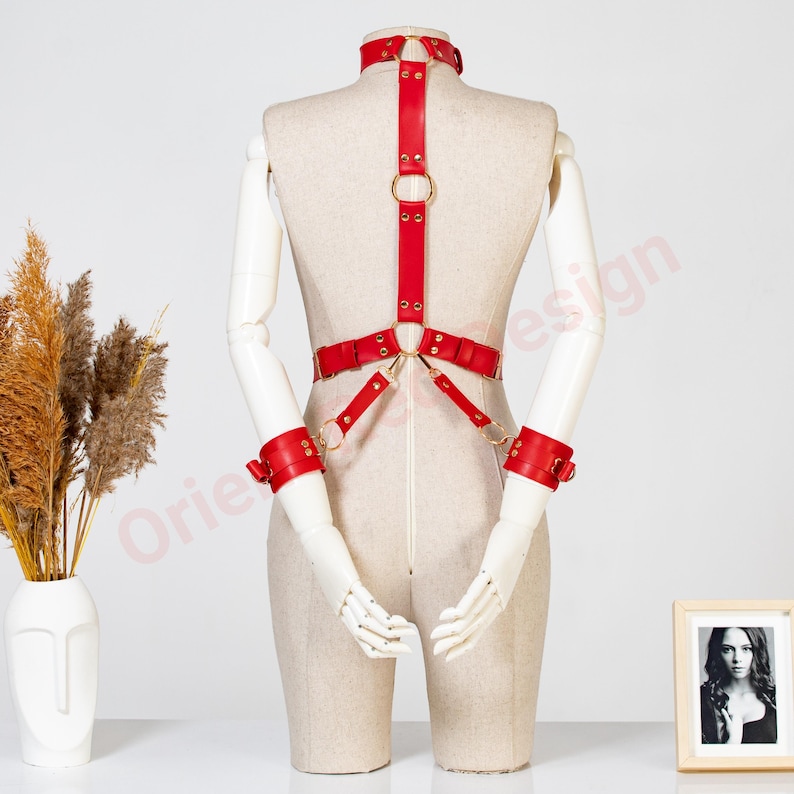 May include: A red leather body harness featuring a neck collar, a vertical strap connecting to a waist belt, and two wrist cuffs. The harness is constructed with gold-toned metal rings, buckles, and rivets, creating a structured design.
