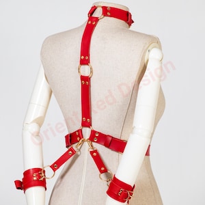 May include: A red leather harness featuring a neck collar, a vertical back strap, and a waist belt connected by gold-toned metal rings and rivets. The harness includes two matching red leather wrist cuffs attached to the waist belt by straps.