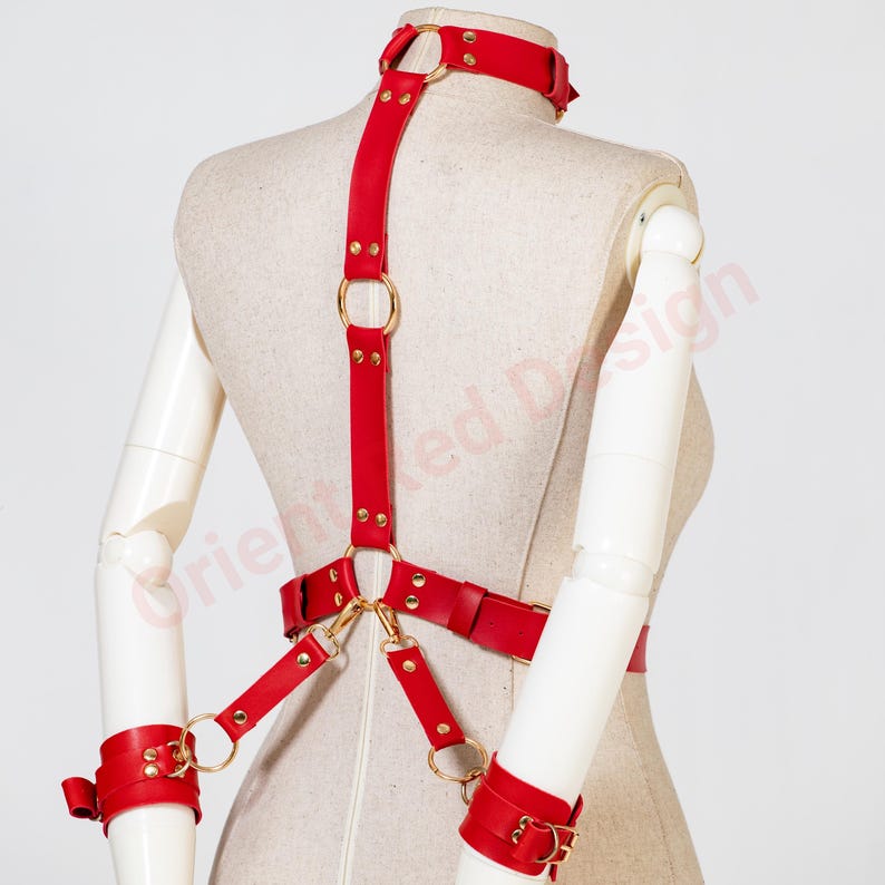 May include: A red leather harness featuring a neck collar, a vertical back strap, and a waist belt connected by gold-toned metal rings and rivets. The harness includes two matching red leather wrist cuffs attached to the waist belt by straps.