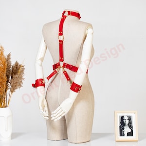 May include: A red leather body harness featuring a neck collar, adjustable straps, and wrist cuffs connected by gold-toned metal rings and hardware. The harness is displayed on a beige mannequin torso with arms positioned behind the back.