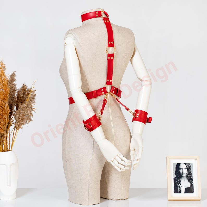May include: A red leather harness set featuring a neck collar, a waist belt, and two wrist cuffs connected by gold-toned metal rings and rivets. The harness is displayed on a mannequin torso, showcasing a structured design with adjustable straps.