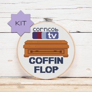 May include: A round embroidery hoop with a cross-stitch design of a brown wooden TV stand with the text "corncob tv" above it and the text "COFFIN FLOP" below it.