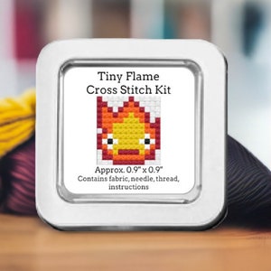 May include: A white metal tin with a clear plastic window showing a cross stitch kit of a cartoon flame. The text on the tin reads "Tiny Flame Cross Stitch Kit" and "Approx. 0.9" x 0.9" Contains fabric, needle, thread, instructions".