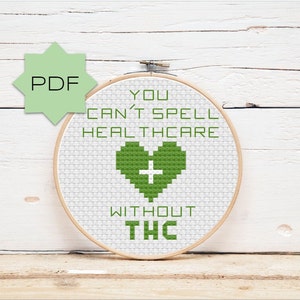 Op de afbeelding: Een kruissteekpatroon in een houten borduurring met de tekst "You can't spell healthcare without THC" en een groen hart met een wit plusteken in het midden.