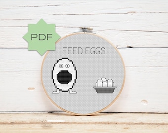 May include: A white embroidery hoop with gray fabric featuring a black and white cross-stitch design of a cartoon egg with googly eyes and the text "FEED EGGS".