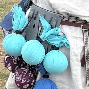 May include: A person wearing a white tunic with a brown leather belt and a variety of fabric pouches attached to it. The pouches are filled with small, round objects, some of which are blue, gray, and red.