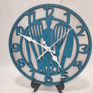 May include: A round, teal-colored kitchen clock with white hands and numbers. The clock face features cut-out silhouettes of kitchen utensils, including a whisk, spatula, and knife. The clock is displayed on a black stand.