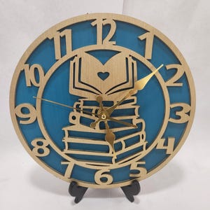 May include: Round wooden clock with a teal blue background and gold-colored hands and numbers. The clock face features a stack of books with an open book on top, and a heart cut out. The clock is displayed on a black stand.