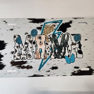 May include: White metal license plate with a black and white cowhide pattern. The word "MAMA" is printed in large letters with a leopard print and a blue lightning bolt.