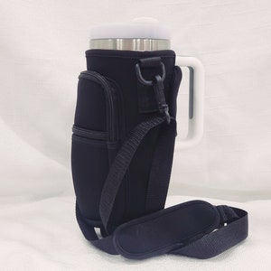May include: Black insulated beverage holder with a zippered pocket, adjustable shoulder strap, and a white handle. The holder is designed to fit a standard-sized tumbler.