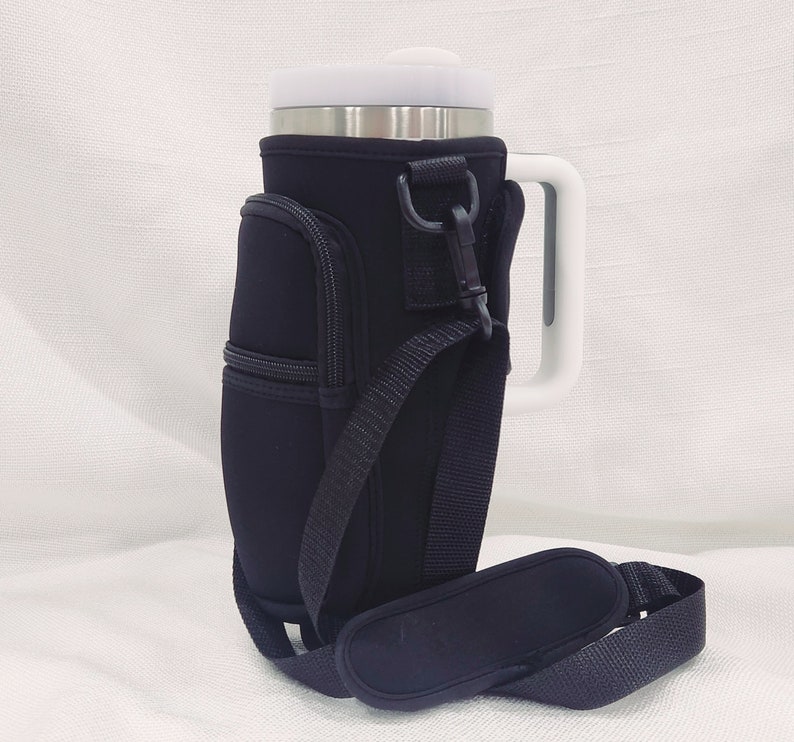 May include: Black insulated beverage holder with a zippered pocket, adjustable shoulder strap, and a white handle. The holder is designed to fit a standard-sized tumbler.