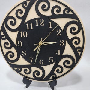 May include: A round wooden clock with a black face and a decorative, swirling design around the perimeter. The clock hands are gold, and the numbers are in a classic font. The clock is displayed on a black stand.