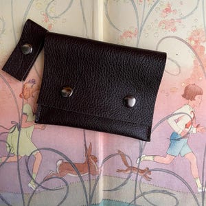 May include: A dark brown leather wallet with a snap closure and a matching strap. The wallet is rectangular and has a textured surface. The wallet is placed on top of a book with illustrations of children and animals.