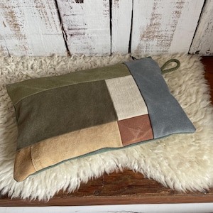 May include: A rectangular patchwork pillow featuring various earth-tone fabrics, including olive green, tan, and brown. The pillow has a loop handle and is placed on a fluffy, white rug against a rustic, white-painted wooden backdrop.