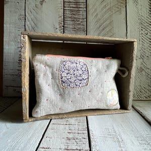 May include: A rectangular beige linen zipper pouch featuring a coral-colored zipper, a central square patch with a dark blue floral pattern, and a smaller circular patch with a light floral print, accented by small red embroidered stitches.