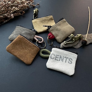 May include: A collection of small, rectangular canvas coin pouches in various colors including tan, gray, brown, and off-white. Each pouch features a black zipper closure, a metal key ring, and a short cord loop. One off-white pouch has the word cents printed on it.