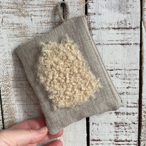 May include: A small, beige linen pouch with a cream-colored, textured wool accent. The pouch has a zipper closure and a small loop for hanging. The background is a distressed white wooden surface.