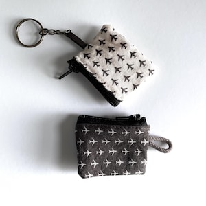 May include: Two small square fabric pouches with zipper closures. The top pouch is cream-colored with a black airplane pattern and a metal keyring attached. The bottom pouch is dark gray with a white airplane pattern and a small fabric loop.