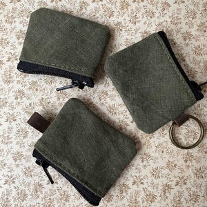 May include: Three olive green canvas coin purses with black zippers are arranged on a floral patterned surface. One purse has a brown leather tab, and another has a metal ring attached. The purses are rectangular with rounded corners.