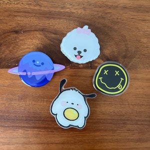 May include: Four colorful pop sockets with cartoon designs. Includes a blue planet with purple rings, a white dog with a pink bow, a yellow smiley face with crossed-out eyes, and a white egg with a yellow yolk.