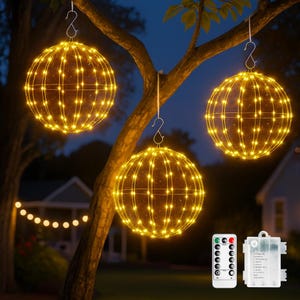 May include: Three illuminated sphere-shaped decorative lights hanging from a tree branch. Each sphere is covered in small, warm yellow lights. A remote control and battery pack are visible. The scene is set outdoors at dusk.