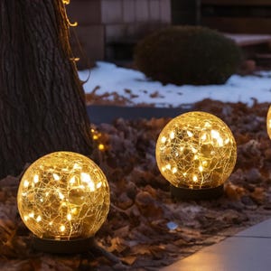 May include: Two illuminated glass globe lights with a crackled texture, filled with warm white fairy lights. The spherical lights sit on black bases, placed outdoors among fallen leaves and snow, creating a cozy ambiance.
