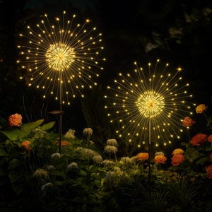 May include: Two outdoor decorative lights with warm white LED lights, resembling fireworks, are displayed in a garden setting. The lights are mounted on black poles and surrounded by green foliage and orange flowers, creating a festive ambiance.