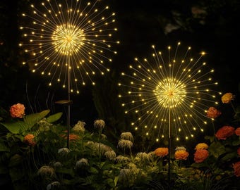 May include: Two outdoor decorative lights with warm white LED lights, resembling fireworks, are displayed in a garden setting. The lights are mounted on black poles and surrounded by green foliage and orange flowers, creating a festive ambiance.