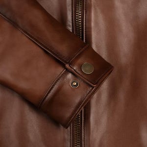 May include: Close-up of a brown leather jacket with a zipper closure and buttoned cuffs. The jacket has a smooth, textured surface.