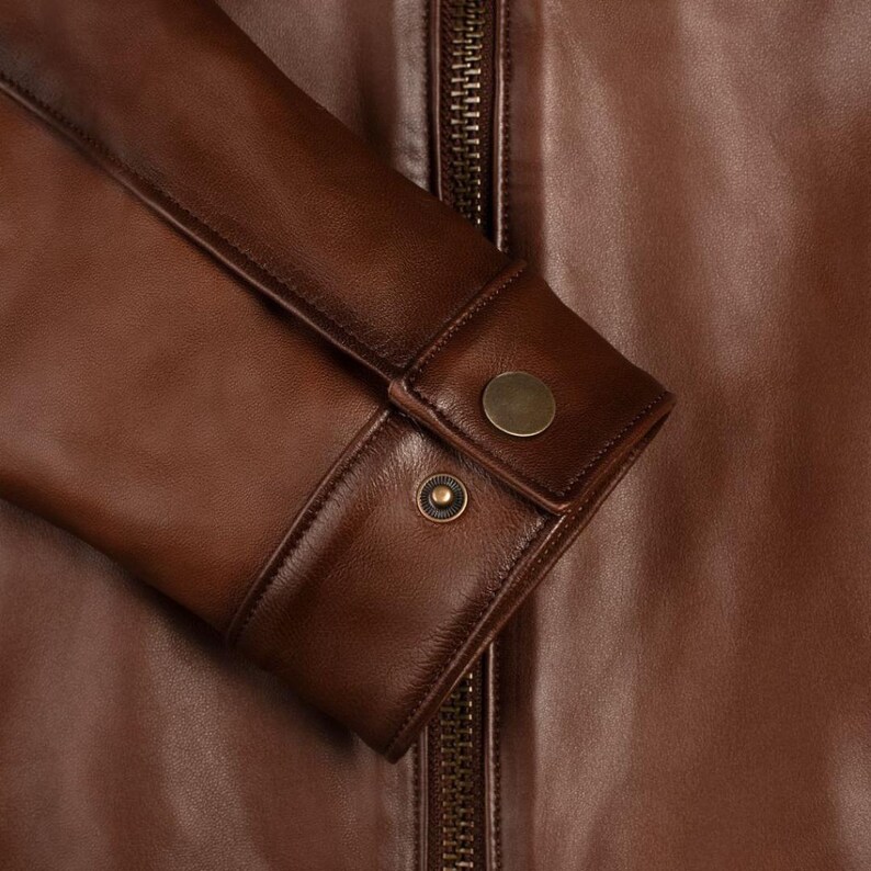 May include: Close-up of a brown leather jacket with a zipper closure and buttoned cuffs. The jacket has a smooth, textured surface.