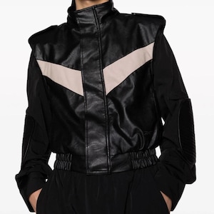 May include: A black leather jacket with a white chevron design on the front. The jacket has a high collar and a zipper closure. The sleeves are long and have black elbow pads.