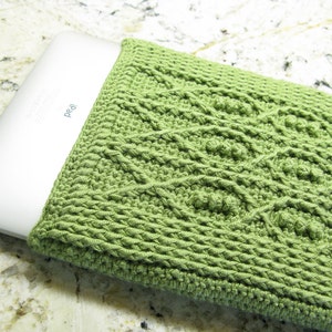 May include: A green crocheted laptop sleeve with a textured pattern. The sleeve is covering a white laptop with the word "iPad" printed on it.