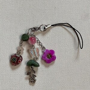 May include: A decorative charm with a mix of beads and charms. It features a heart-shaped bead with a floral design, a pink flower, a butterfly, and other beads in pink, green, and clear. It's attached to a silver clasp and a black cord. The charm is approximately 10 cm long.