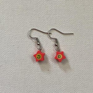 May include: A pair of earrings with red star-shaped charms, yellow centres, and a smiling face. The earrings have silver-coloured hooks.