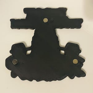 Pu&ograve; includere: Un oggetto nero, piatto e dalla forma personalizzata con una superficie ruvida e tre magneti circolari argentati incorporati sul retro. L'oggetto presenta una sezione superiore larga e una sezione inferiore pi&ugrave; ampia e sagomata.