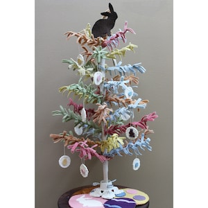 May include: A pastel-colored Easter tree with a black bunny topper and hanging egg and bunny ornaments. The tree has a white trunk and sits on a small, round table with felt Easter decorations. The tree's branches are in shades of pink, blue, yellow, and brown.